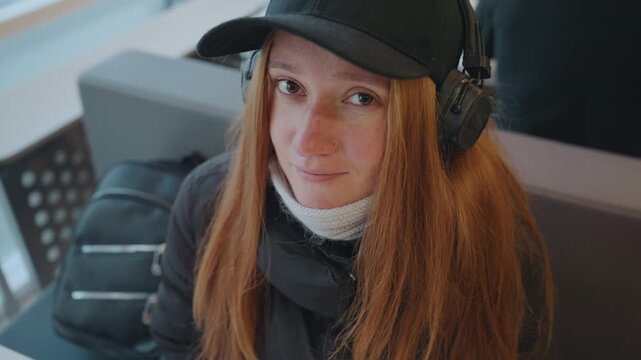 White woman with headphones in cafe, redhead commuter sits by window with backpack, glances around pensive, subtle smiles and freckled cheeks, bundled in coat and cap, urban travel tension, roles