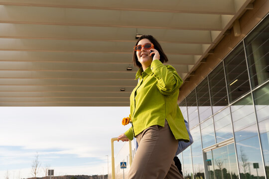 Woman talks on phone while walking with suitcase outside building