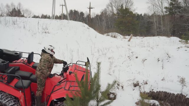 Camouflage rider on red atv ready at snowy bank rider adjusts stance engine idling anticipation before creek crossing powertrain hum poles and pine trees in background cold overcast light cautious