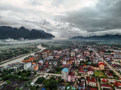 Aerial view of the town of Vang Vieng with the Nam Song River winding past limestone karst mountains under a misty, overcast sky in Vang Vieng, Vientiane Province, Laos.