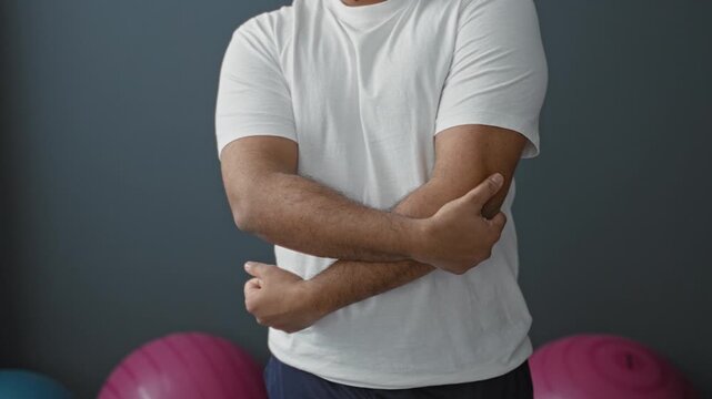 Man holding elbow with crossed arms in studio with exercise balls and gray wall wearing white tee; recovery calm.