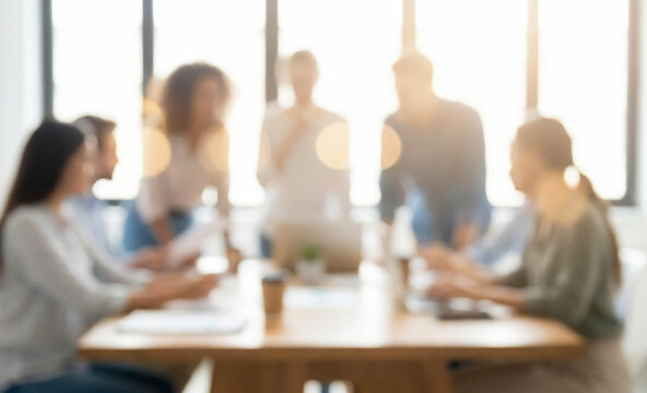 A blurred view of a diverse group of people engaged in a collaborative meeting around a wooden table, with soft natural light illuminating the space.