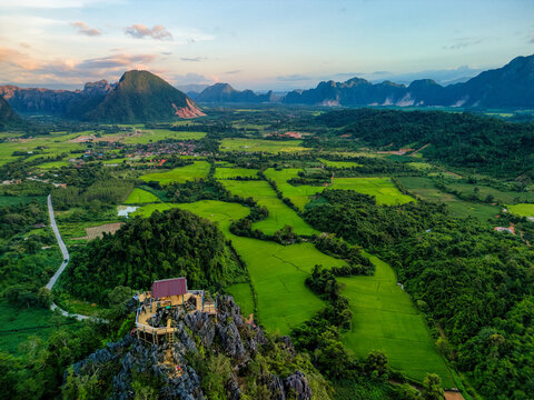 Aerial view of the Nam Xay viewpoint on a limestone peak overlooking lush green rice fields and karst mountains in Vang Vieng, Vientiane Province, Laos.