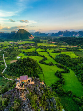 Aerial view of a wooden viewpoint on a rocky peak with people overlooking lush green rice paddies and karst mountains under a soft sunset sky in Vang Vieng, Vientiane Province, Laos.
