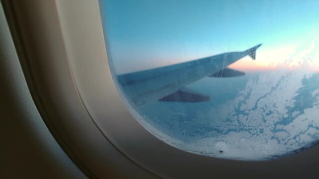 Airplane wing visible through airplane window with condensation, showcasing the changing sky colors during flight over clouds at sunset