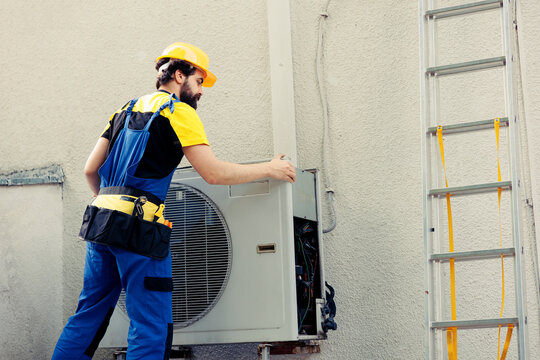 Skilled wireman starting work on malfunctioning air conditioner, using electric drill to take apart condenser coil panel. Experienced serviceman dismantling hvac system to check for faulty wires