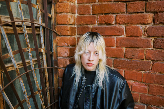 Young Person With Light Hair Poses by Brick Wall With Metal Grating