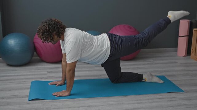 Young indian man on blue yoga mat performing leg extension and core stabilization exercise in studio; focus balance.