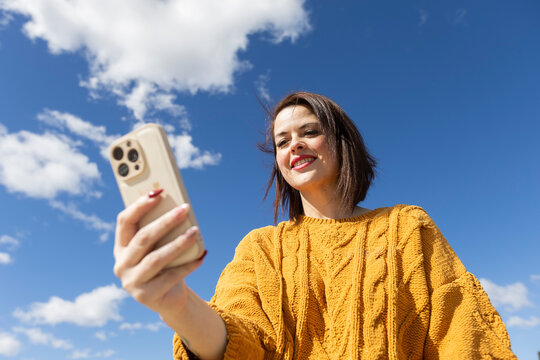 Smiling woman holding smartphone in sunlight over blue sky