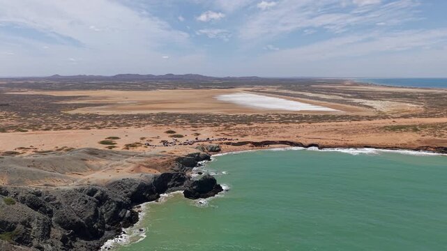 Sweeping aerial drone footage reveals Cabo de la Vela&rsquo;s turquoise bay, rugged cliffs, and golden desert contrasting with white salt flats under bright natural daylight in La Guajira.
