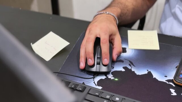 Close-up of a hand using a computer mouse on an office desk with sticky notes, smartphone, and monitor in an indoor workspace under soft lighting.