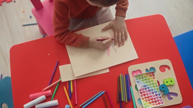 Boy tracing a hand and holding a red crayon while coloring on paper at a red kids table in a studio with stickers and colored pencils; concentration.