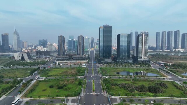 Wide view of developing district with symmetrical boulevard leading toward modern skyline and bridge, surrounded by green plots under cloudy daylight.