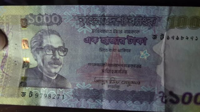 Front view of one thousand bangladeshi taka note being held by hands against dark surface in close up cinematic footage showing portrait of sheikh mujibur rahman and intricate currency designs.