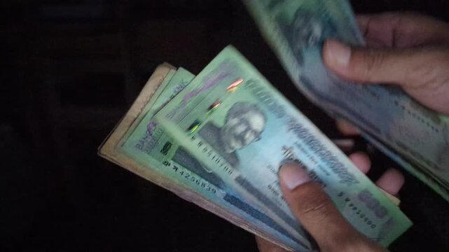 Fingers meticulously count stack of Bangladeshi taka banknotes displaying portrait of Sheikh Mujibur Rahman in low light closeup showing currency motion.