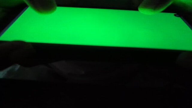Hands using green screen smartphone in dark environment for mobile app prototyping and interface testing with chroma key display on electronic device screen for digital content production work.