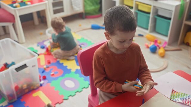 Boy holding colored pencils and drawing at a small table while younger brother plays on a foam mat in a playroom building; concentration sibling bond.