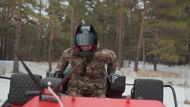 Snowy forest ATV rider gear check. Individual dressed in black preparing ATV amidst snowy trees. Caucasian individual in camouflage attire proceeding with ATV check in winter forest environment