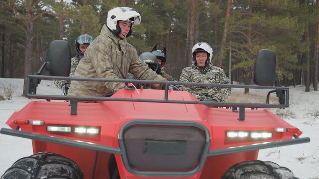 Caucasian rider stands on red atv with headlights on, driver scanning snowy pine trail, two helmeted passengers seated in camouflage, focused posture and engine rumble signal readiness for extreme