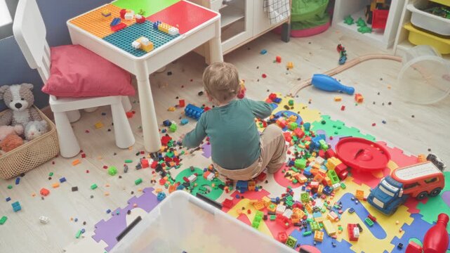 Boy sitting on foam puzzle mat reaching for scattered toy blocks and red toy truck in a cluttered play area inside a building; playful curiosity.
