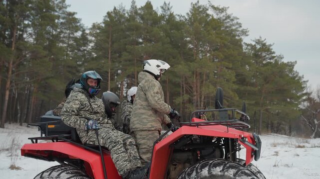 Red atv in snowy forest caucasian duo in camouflage suits and helmets riding across pine clearing, tires throwing snow, passenger waving, guide role suggested, playful camaraderie, offroad winter