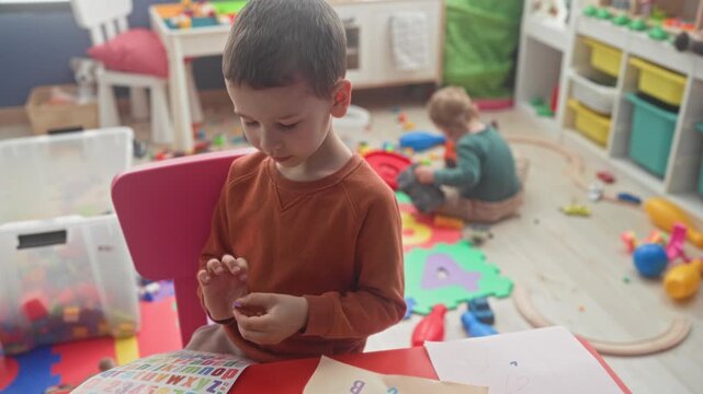 Boy studies sticker sheet, fingers peeling sticker at a play table in a building playroom with a younger brother on the floor; concentration sibling bond.