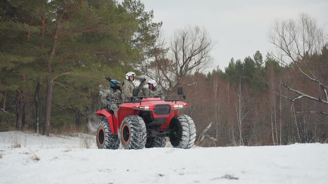 Three riders on red atv winter, group wearing helmets and camo jackets, heavy tread tires carving through packed snow, machine charging across forest clearing, energetic camaraderie and teamwork,
