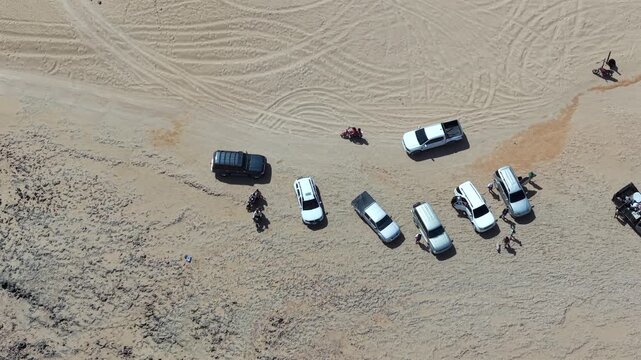 Drone video capturing parked vehicles and people on Playa Arcoiris in Cabo de la Vela, La Guajira, Colombia. Bright sunlight casts crisp shadows on rugged sand in this dramatic aerial landscape.