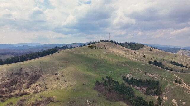 Aerial drone view of Toaca Hill in Gura Humorului Romania featuring the large hillside sign and rolling grassy landscape with forest patches under cloudy sky