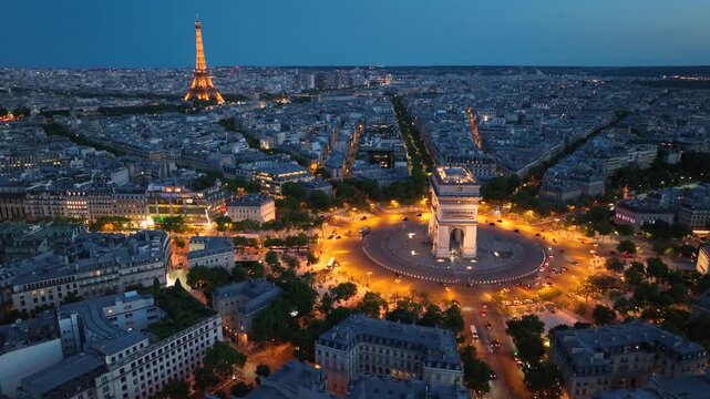 Aerial night view of the famous landmark Triumphal Arch or Arc de Triomphe and Eiffel Tower surrounded by busy streets and lush greenery in the heart of the city Paris, France. Drone orbiting flight