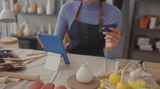 Woman holding creditcard by tablet stand and sculpting tools on table in artisan studio indoors; small business confidence.