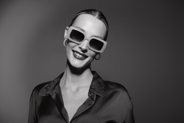 Obraz premium Smiling Caucasian woman with pale skin wearing oversized sunglasses and hoop earrings poses confidently in a dark satin blouse on grey background. Fashion and style concept.