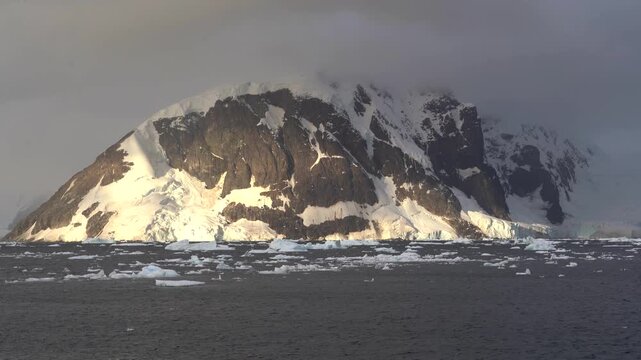 Steady wide-angle shot of a massive, snow-dusted mountain on Danco Island illuminated by low golden-hour sun, featuring brash ice floating in the Errera Channel