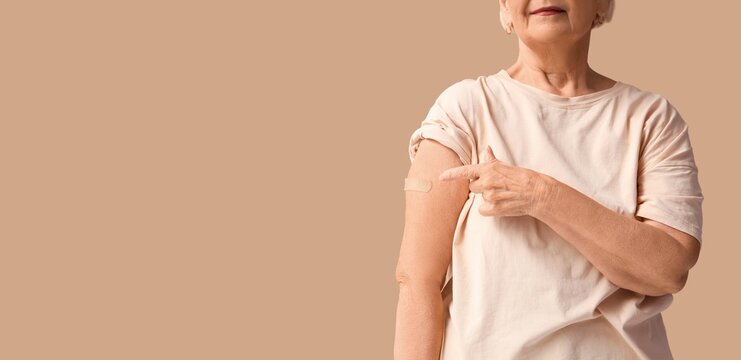 Senior woman pointing at medical patch on her arm after vaccination against beige background with space for text