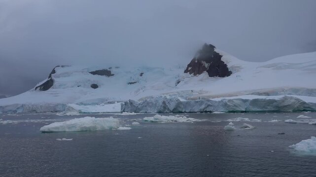 Cinematic POV shot moving slowly past towering blue-tinted ice walls and floating brash ice at the base of snow-capped mountains in the Antarctic Peninsula