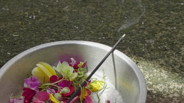 A close up of an aluminum plate filled with flowers and burning incense, releasing soft smoke. Used in Balinese Hindu prayer rituals, capturing sacred atmosphere and cultural spirituality.