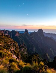 Fototapeta premium A majestic mountain range is silhouetted against a pastel sky with a visible crescent moon. The foreground features grassy slopes