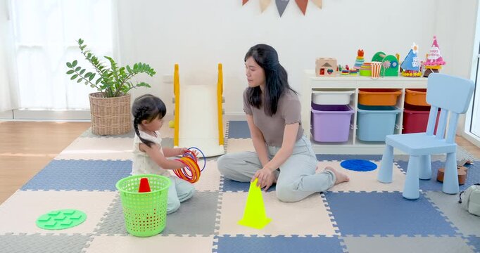 Asian single mother sits with preschool daughter in home playroom, guiding ring toss game and motor skill practice on foam mat while little girl reaches for colorful toy during learning time.