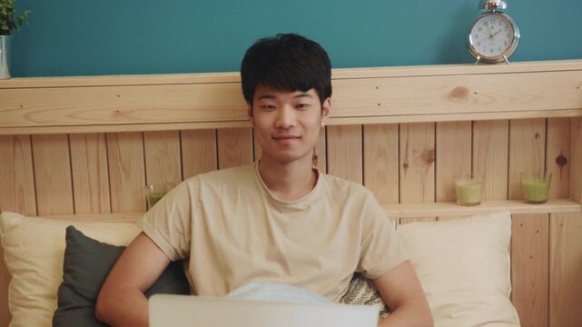 Smiling man using laptop in warm bedroom, Content Asian man engaged with computer in inviting bedroom environment