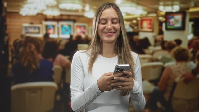 Young woman holding smartphone and smiling while looking aside in a building waiting area crowded with seated people; contentment social connection.