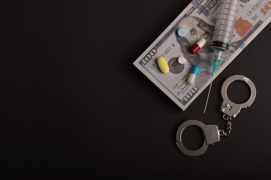 Syringe, pills, handcuffs, and dollar banknotes on dark background. Concept of drug trafficking, illegal substances, financial crime, and law enforcement.