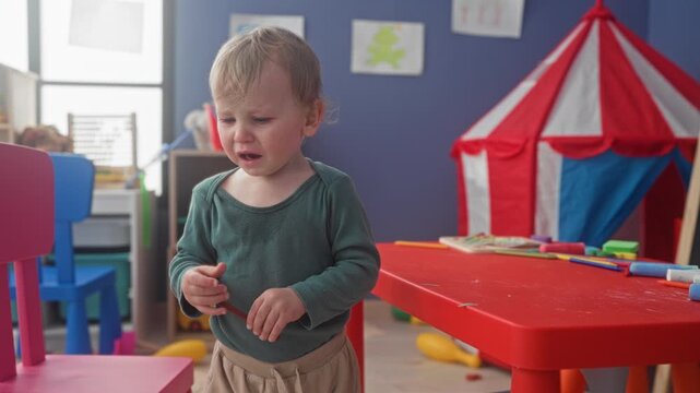 Boy crying, hands gripping crayon at red table beside play tent in a daycare playroom inside building; tantrum distress.