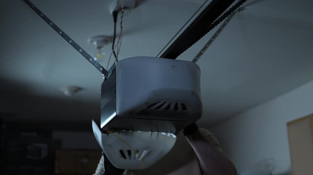 ServiceMan repairing garage door opener. Technician checks wires on ceiling mounted motor in residential garage. Close up