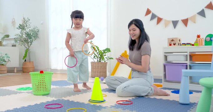Asian single mother watches preschool daughter prepare ring toss in home playroom, supporting balance, coordination, and problem solving through playful learning with cone target and colorful hoops.