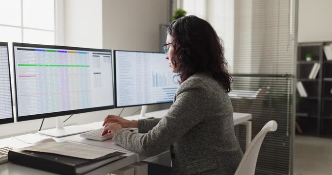 Woman analyst accountant at office computer analyzing spreadsheet. She checks figures on multiple monitors for accuracy and timely corporate reporting. Clear concept of financial data analysis.
