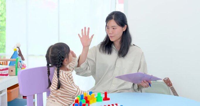 Asian single mother gives preschool daughter high five after successful block activity, celebrating progress, confidence, parent child bonding, motivation, and positive learning growth in playroom.