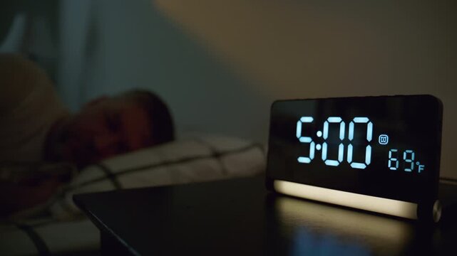 Man turning off alarm clock at 5,00 in bedroom. Early morning wake up moment with digital clock beside bed, 