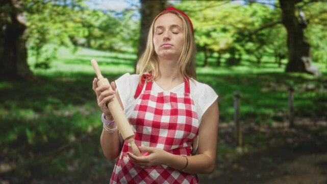 Woman chef, blonde, wearing red gingham apron and headband, holding a wooden rolling pin, tilting head and flicking fingers in a forest setting; boredom.