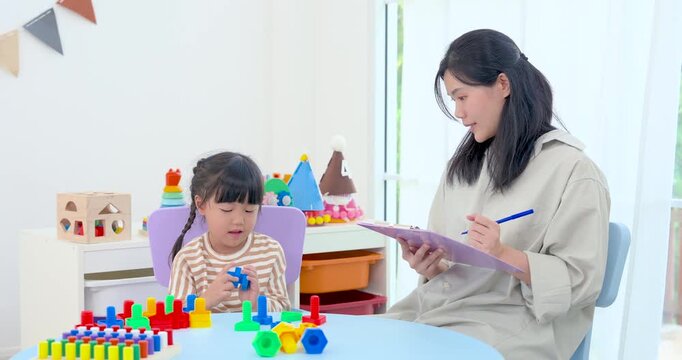 Asian mom checks notes on tablet while preschool daughter concentrates on fitting blue block piece at table, building shape recognition, persistence, fine motor control, and playful learning indoors.
