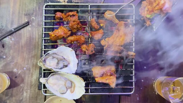 Overhead view of sizzling seafood barbecue in Hoi An, Vietnam, with oysters, grilled meat, smoke, and glowing coals creating a vibrant street-food cooking scene.